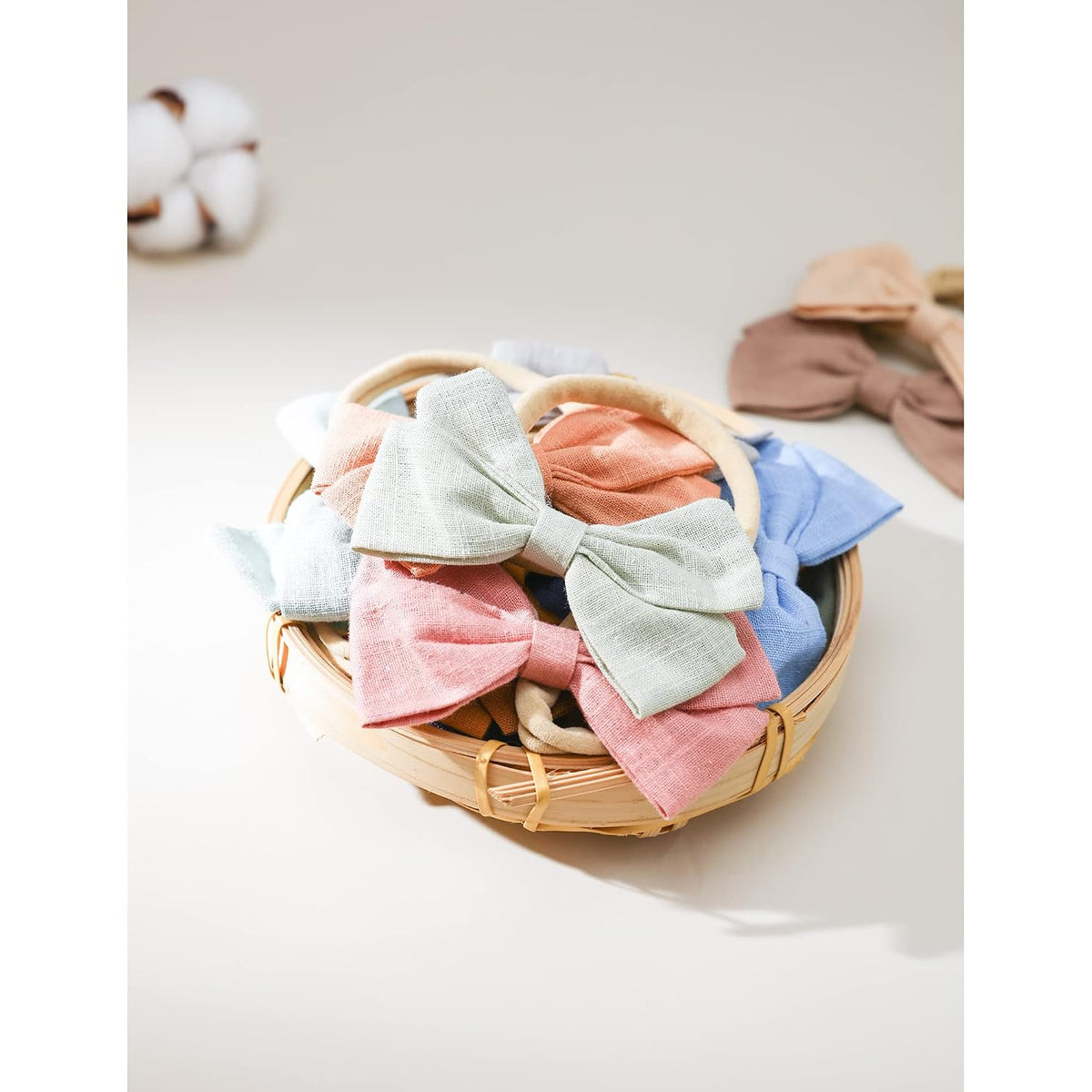 A round woven bamboo basket filled with fabric bow headbands in soft pastel colors including pink, sage green, coral, and blue. Two additional bow headbands in tan and brown rest beside the basket. A cotton boll is visible in the upper left corner.