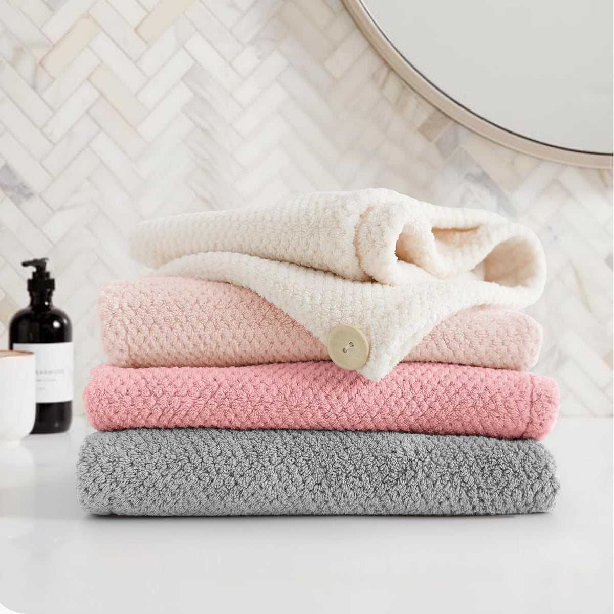 Stack of four textured microfiber hair towel wraps in gray, pink, light pink, and cream, folded and stacked on a white surface. The cream towel on top is loosely wrapped and features a small button closure. Bathroom setting with herringbone tile backdrop.