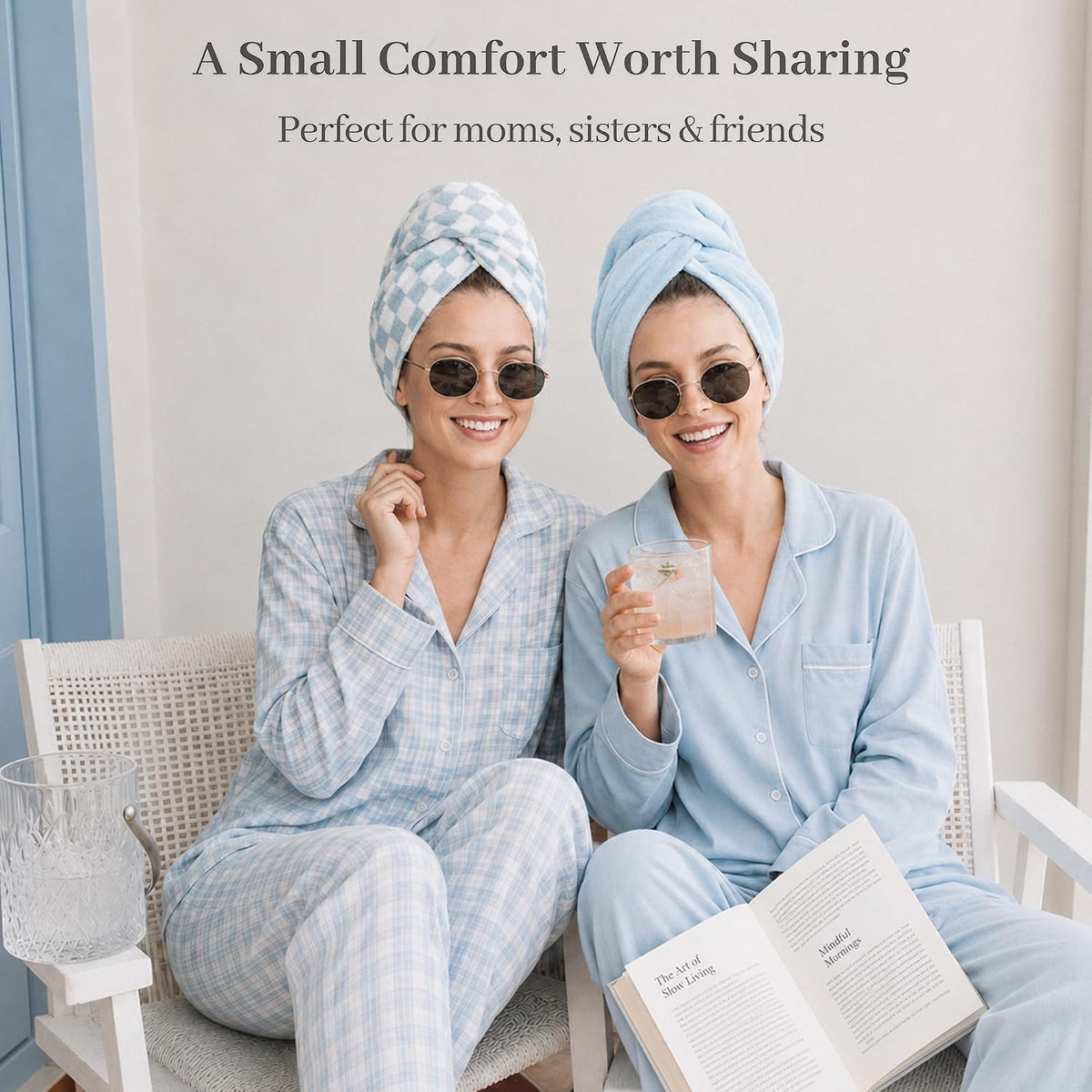 Two smiling women in blue pajamas and round sunglasses sit together, each wearing a hair towel turban. One wears a blue checkered wrap, the other a solid light blue wrap. Text reads 'A Small Comfort Worth Sharing - Perfect for moms, sisters & friends.'