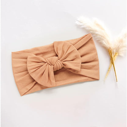 Soft fabric baby headband in warm caramel tan, laid flat with a large knotted bow at center. Shown on a white surface beside a dried pampas grass stem.