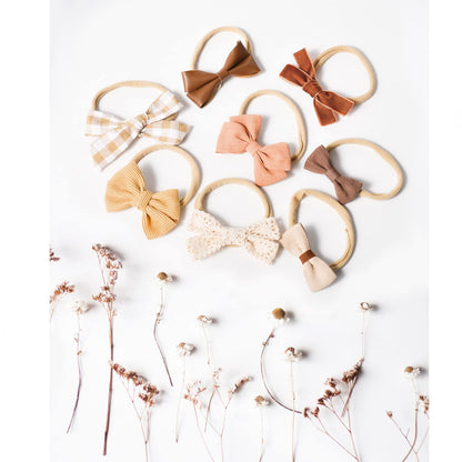 Eight baby bow headbands on soft nylon elastic bands, arranged on a white surface. Bows vary in fabric and color: gingham, caramel leather-look, rust, blush linen, mauve, mustard ribbed, cream lace, and beige waffle knit.