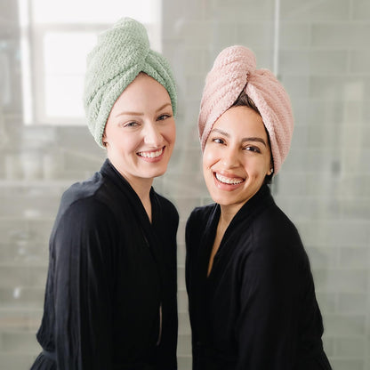 Two smiling women wearing twisted hair towel wraps, one in sage green and one in dusty pink, both dressed in black robes, shown from the front in a bathroom setting.