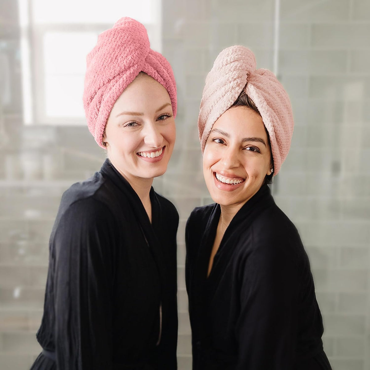 Two smiling women in black robes wearing twisted hair towel wraps in a bathroom setting. One wrap is bright pink, the other is dusty rose/mauve, both with a textured, looped fabric and knotted twist at the top.