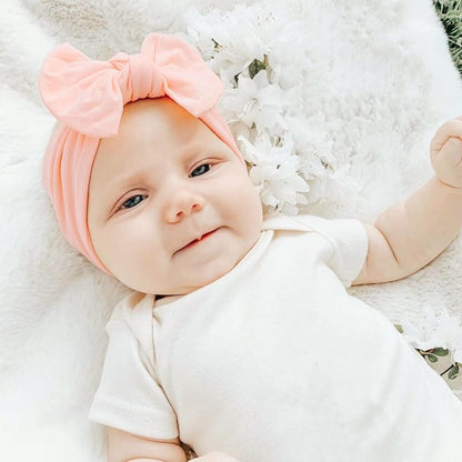 Baby wearing a soft peach-pink nylon turban-style headband with a large bow on top, lying on a white fluffy blanket with white flowers nearby, dressed in a cream onesie.