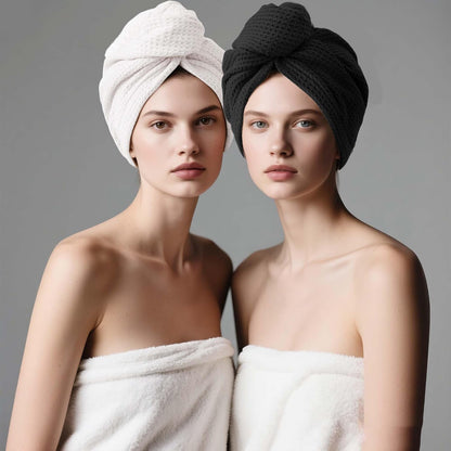 Two models each wearing a twisted hair towel turban wrap, one in white waffle-weave fabric and one in black waffle-weave fabric, both draped in white towels against a gray background.