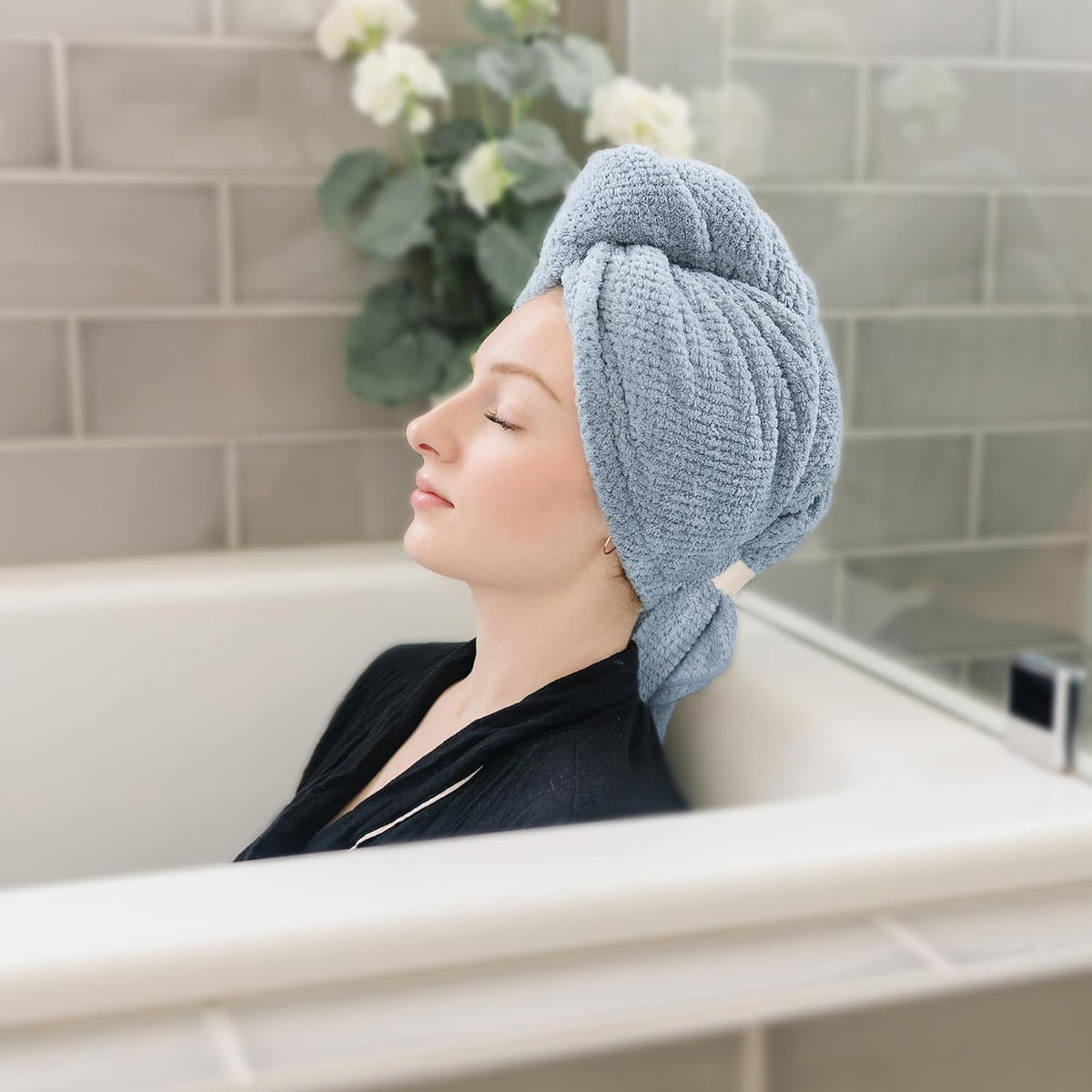 Woman relaxing in a bathtub wearing a light blue textured hair towel wrapped in a turban style on her head, eyes closed, in a tiled bathroom with white flowers in the background.