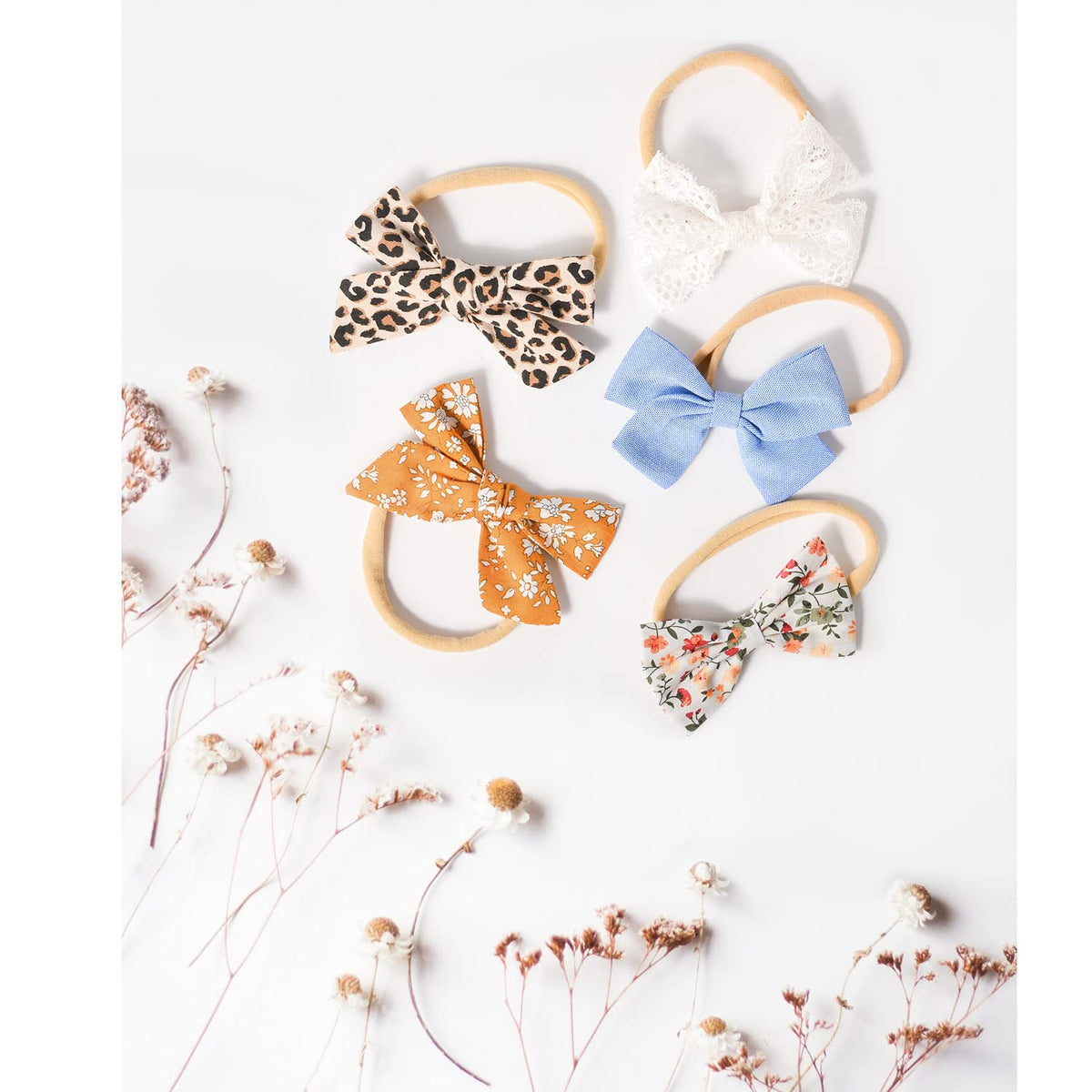 Five fabric bow headbands on thin nylon elastic bands arranged on a white surface: leopard print, white lace, light blue solid, golden floral, and multicolor floral. Dried wildflowers accent the lower left.