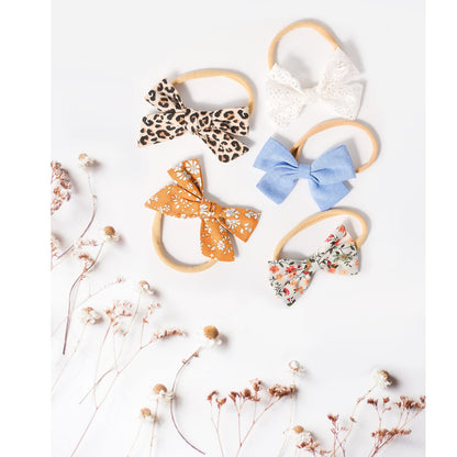 Five fabric bow headbands on thin nylon elastic bands arranged on a white surface: leopard print, white lace, light blue solid, golden floral, and multicolor floral. Dried wildflowers accent the lower left.