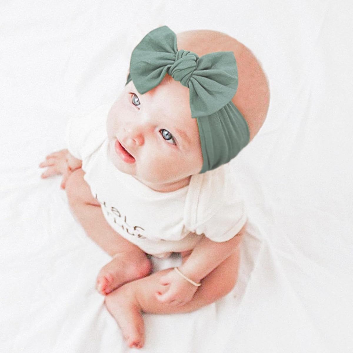 Baby wearing a muted green nylon headband with a large bow tied at the front, shown on a white background. The stretchy band wraps snugly around the infant's head, demonstrating fit and style.