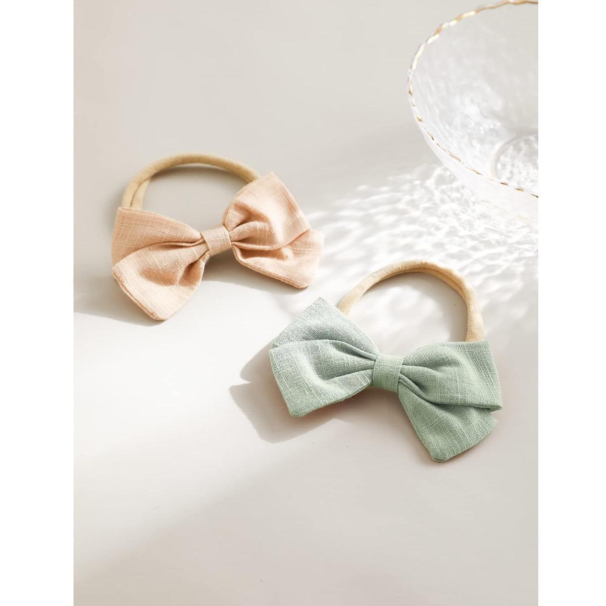 Two fabric bow hair ties on elastic bands laid flat on a light surface. One bow is soft peach and one is sage green, both with a textured linen-like weave. A decorative dish is partially visible in the upper right corner.