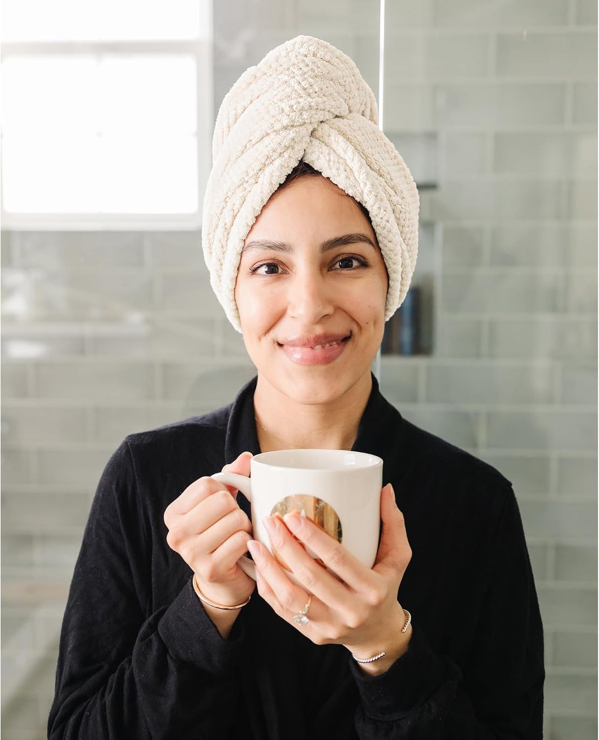 Woman wearing a cream-colored textured hair towel wrapped in a turban style on her head, smiling and holding a white mug. She wears a black robe, shown from the chest up against a gray tile background.