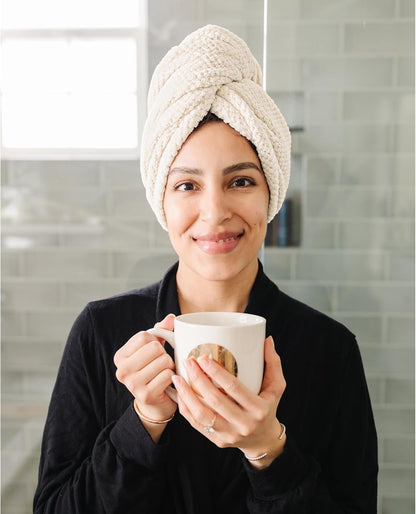 Woman wearing a cream-colored textured hair towel wrapped in a turban style on her head, smiling and holding a white mug. She wears a black robe, shown from the chest up against a gray tile background.