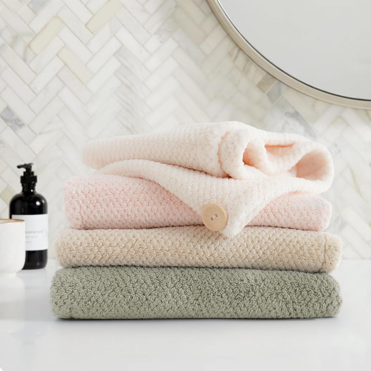 Stack of four folded textured towels in light pink, blush pink, cream, and sage green, displayed on a white surface with a black soap dispenser and round mirror visible in the background.