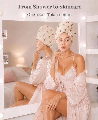 Woman wearing a cream hair towel turban with small red floral print, seated in front of a lit vanity mirror in a pink satin robe. Text reads 'From Shower to Skincare - One towel. Total comfort.'