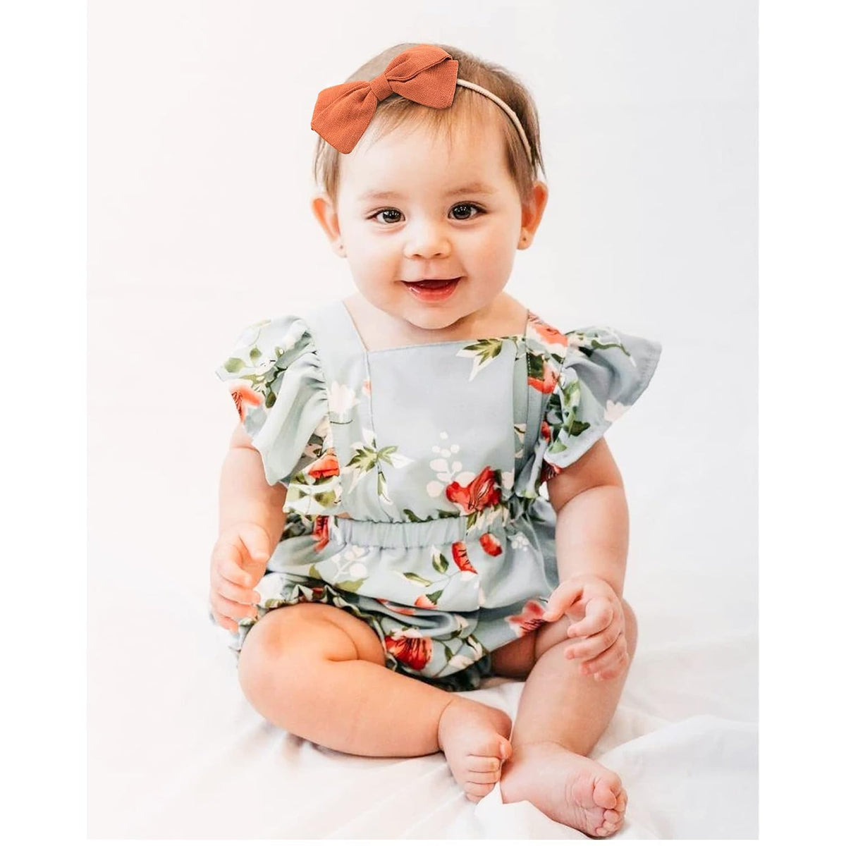 Baby girl wearing a rust-orange linen bow headband on a thin elastic band, paired with a light blue floral romper with ruffle sleeves. She is seated and smiling against a white background.