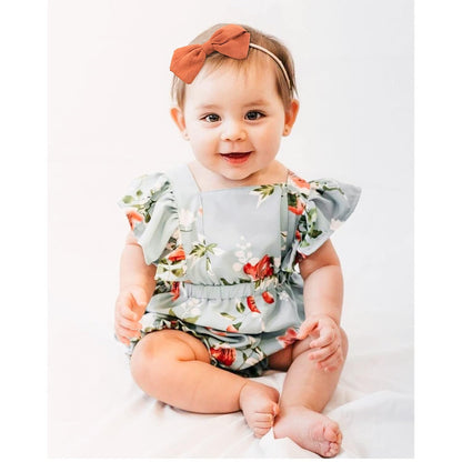 Baby girl wearing a rust-orange linen bow headband on a thin elastic band, paired with a light blue floral romper with ruffle sleeves. She is seated and smiling against a white background.