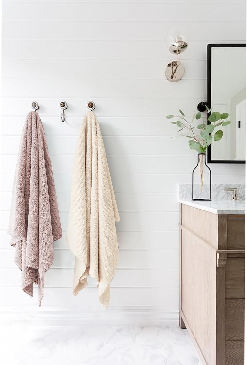 Two large textured towels hanging on silver wall hooks in a bright bathroom with shiplap walls. Left towel is dusty mauve/pink, right is cream/beige. Both have a waffle-knit texture and drape to floor length.