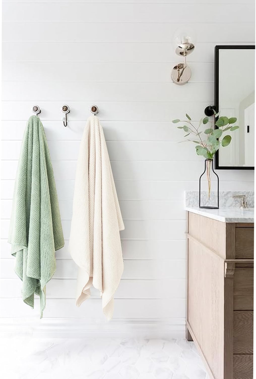 Two large towels hanging on chrome wall hooks in a bright bathroom with shiplap walls. Left towel is sage green with a waffle texture; right towel is cream/off-white. A wood vanity with marble countertop is visible on the right.