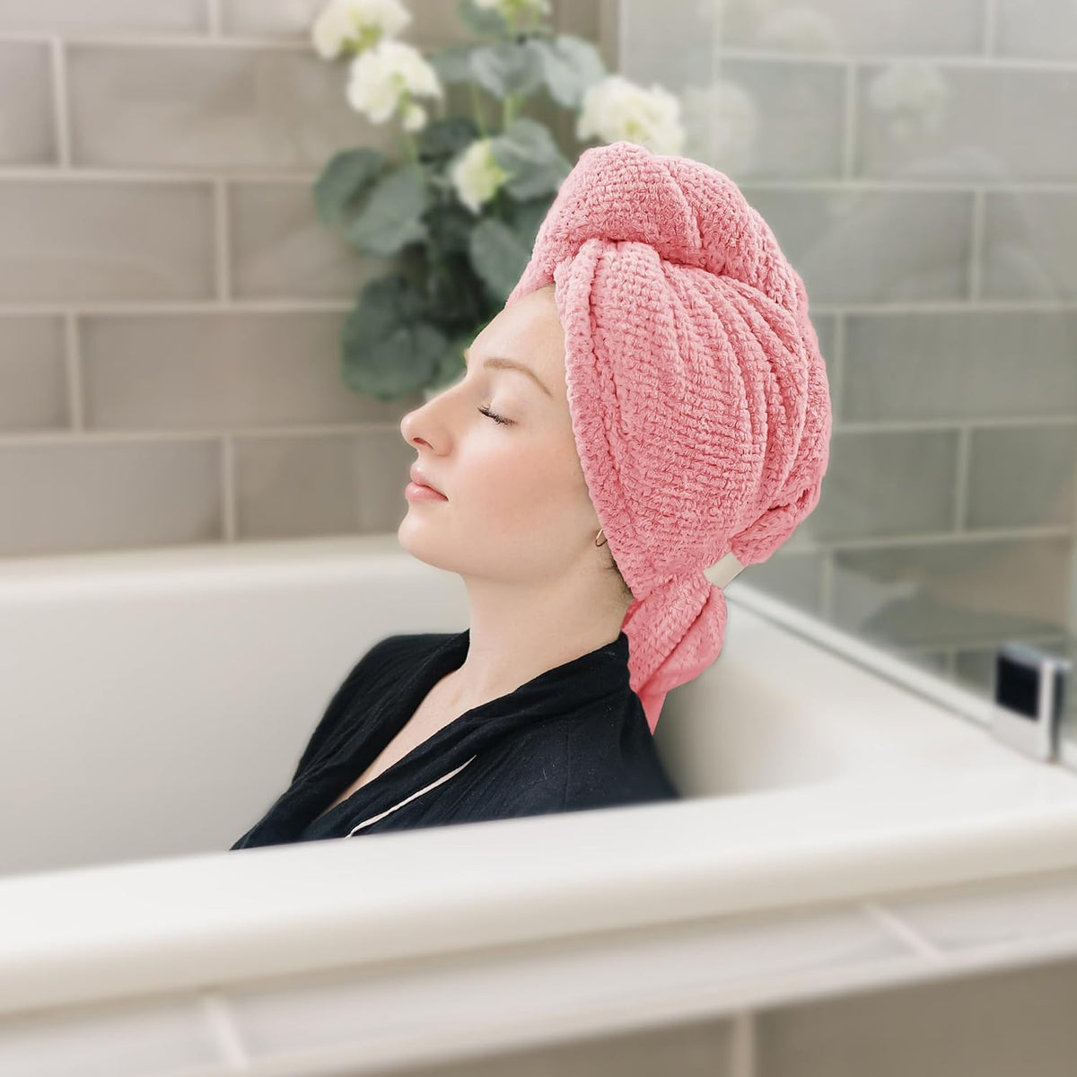 Woman relaxing in a bathtub with eyes closed, wearing a pink textured hair towel wrapped in a turban style on her head. The waffle-weave towel is twisted and knotted at the front. Bathroom tile wall and greenery visible in background.