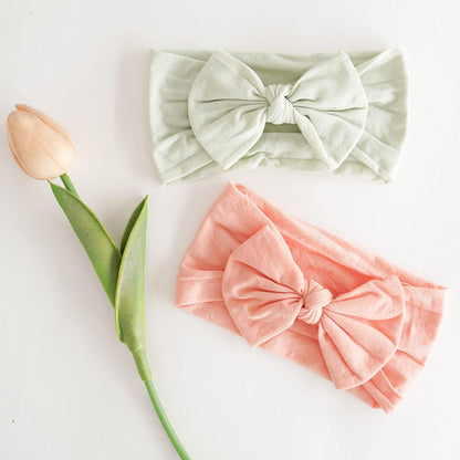 Two soft fabric bow headbands laid flat on a white surface beside a peach tulip. One headband is pale sage green, the other is coral pink. Both feature a large knotted bow at the center.