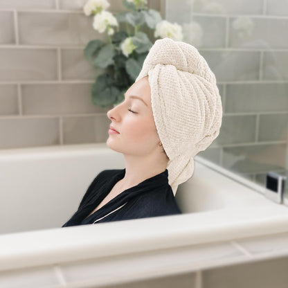 A woman relaxing in a bathtub with eyes closed, wearing a cream-colored textured hair towel wrap twisted and secured on top of her head. Gray tile wall and greenery visible in background.