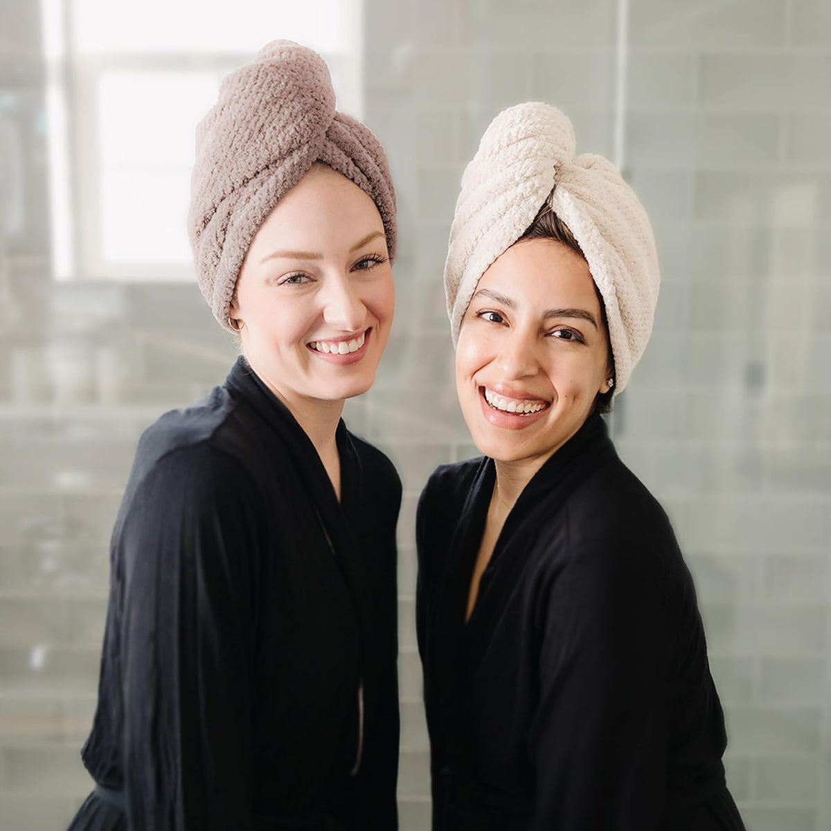 Two smiling women wearing hair towel turbans, both dressed in black robes. One wears a taupe-toned turban, the other a cream-colored turban, both twisted and knotted on top of their heads. Bathroom setting in background.