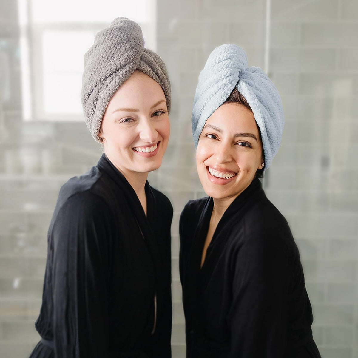 Two smiling women wearing twisted hair towel turbans, one in gray and one in light blue, both dressed in black robes. The towels are wrapped and secured at the top of their heads in a classic turban style.
