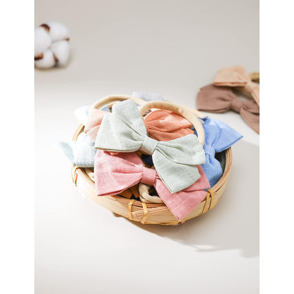 A round woven bamboo basket filled with fabric bow headbands in soft pastel colors including pink, sage green, coral, and blue. Two additional bow headbands in tan and brown rest beside the basket. A cotton boll is visible in the upper left corner.