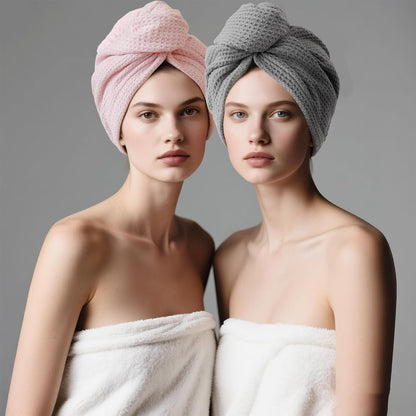 Two models each wearing a twisted hair towel turban wrap, one in light pink and one in gray, both with waffle-weave texture. Models are draped in white towels against a neutral gray background.