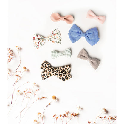 Seven fabric hair bows arranged on a white surface with dried flowers. Styles include solid dusty pink, floral, blue textured, sage green gingham, gray polka dot, and a large leopard print bow.