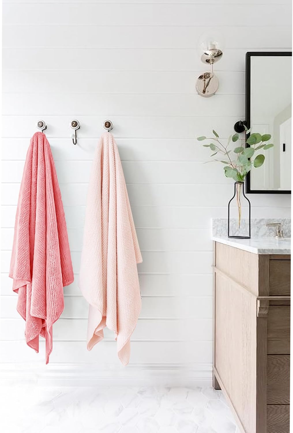 Two large bath towels hanging on silver wall hooks in a bright bathroom. Left towel is coral pink, right is blush pink. Both have a textured waffle-like weave. Light wood vanity and mirror visible on the right.