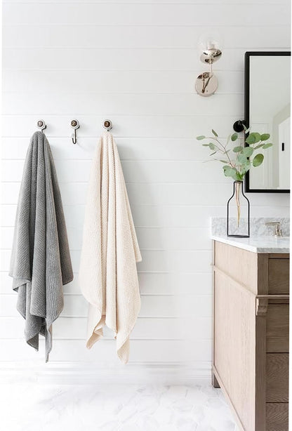Two large textured towels hanging on wall hooks in a bright bathroom — one gray and one cream/beige. Both have a waffle-knit texture and drape to near floor length. A wood vanity with marble countertop is visible to the right.