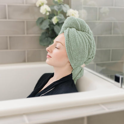 Woman relaxing in a bathtub with eyes closed, wearing a sage green textured hair towel wrapped in a turban style on her head. The towel has a waffle-knit texture and a small loop or button closure visible at the side.