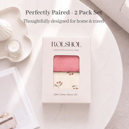 ROLSHOL Premium 100% Cotton Hair Towels 2-pack in a white gift box. Top towel is solid dusty pink; bottom is cream with a cherry print. Box reads 'Soft Cotton, Secure Fit.' Styled on a marble table.