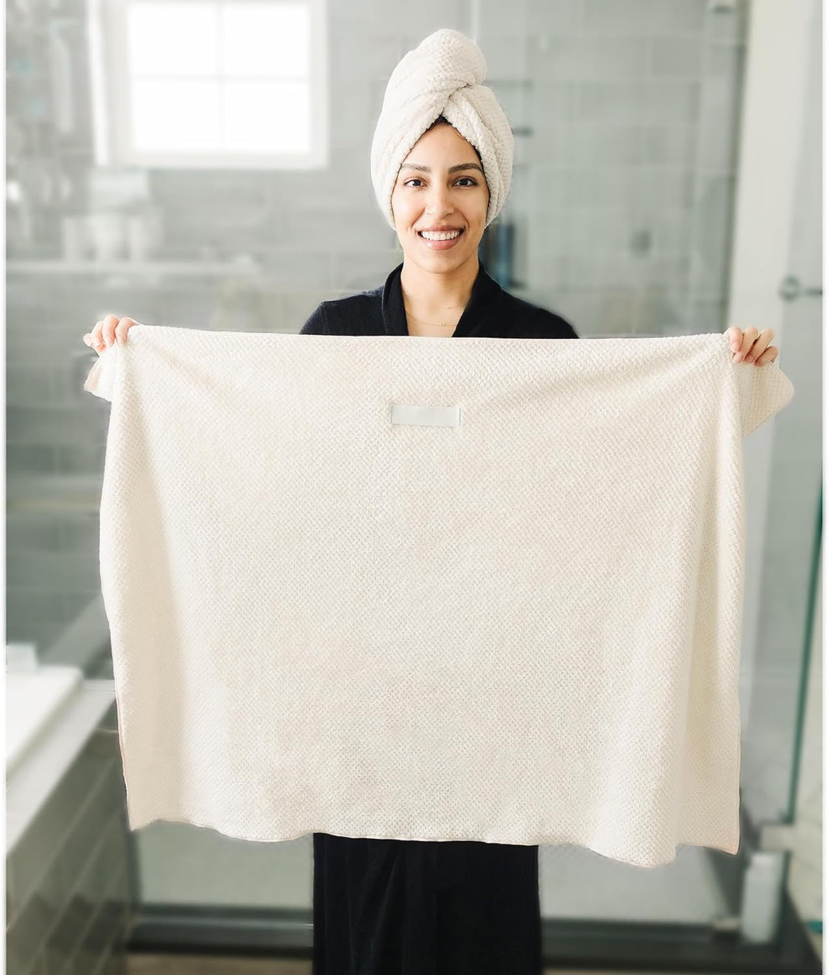 Smiling woman in a black robe holding up a large cream-colored textured bath towel with both hands, displaying its full size. She wears a matching white towel wrapped around her hair. Bathroom setting visible in background.