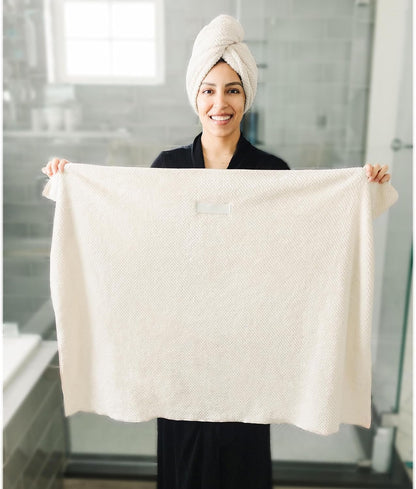Smiling woman in a black robe holding up a large cream-colored textured bath towel with both hands, displaying its full size. She wears a matching white towel wrapped around her hair. Bathroom setting visible in background.