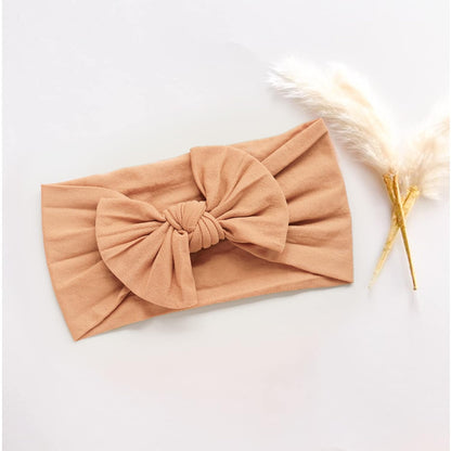 Soft fabric baby headband in warm caramel tan, laid flat on a white surface. Features a large gathered bow knot at center. A dried pampas grass stem is styled alongside it as decoration.