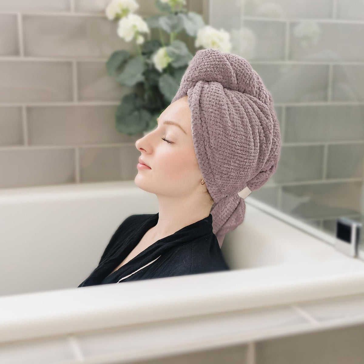 Woman relaxing in a bathtub with eyes closed, wearing a mauve waffle-textured hair towel wrapped in a turban style on her head. A small light-colored tag is visible on the side of the wrap.