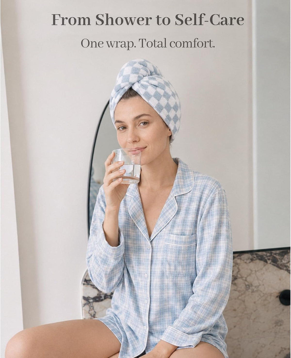 Woman wearing a blue and white checkered hair towel turban on her head, sipping water from a glass. She wears a light blue plaid button-up shirt. Text reads 'From Shower to Self-Care. One wrap. Total comfort.'