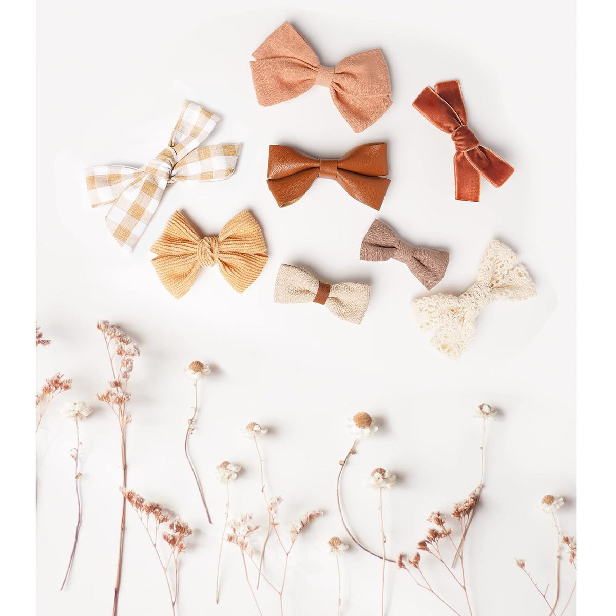 Eight fabric hair bows in autumn tones arranged on a white surface: gingham, blush linen, rust velvet, caramel faux leather, mustard ribbed knit, cream woven, taupe linen, and ivory lace. Dried wildflowers accent the bottom.