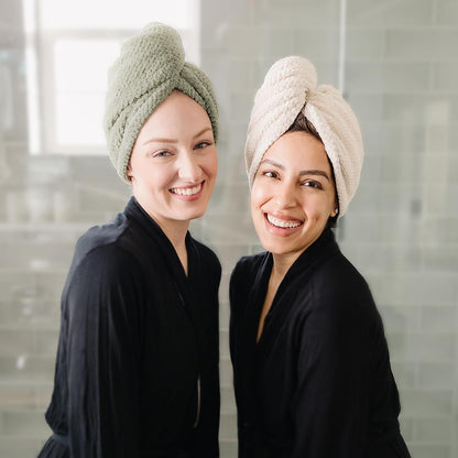 Two smiling women wearing twisted hair towel turbans, one in sage green and one in cream/beige, both dressed in black robes. The textured microfiber turbans are knotted at the top and worn in a bathroom setting.