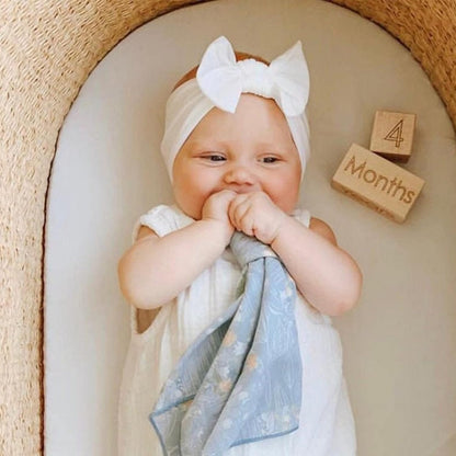 Baby wearing a large white bow headband, sitting in an arched cream-colored seat, holding a light blue floral cloth. Wooden milestone blocks beside her read '4 Months.' She wears a white dress.