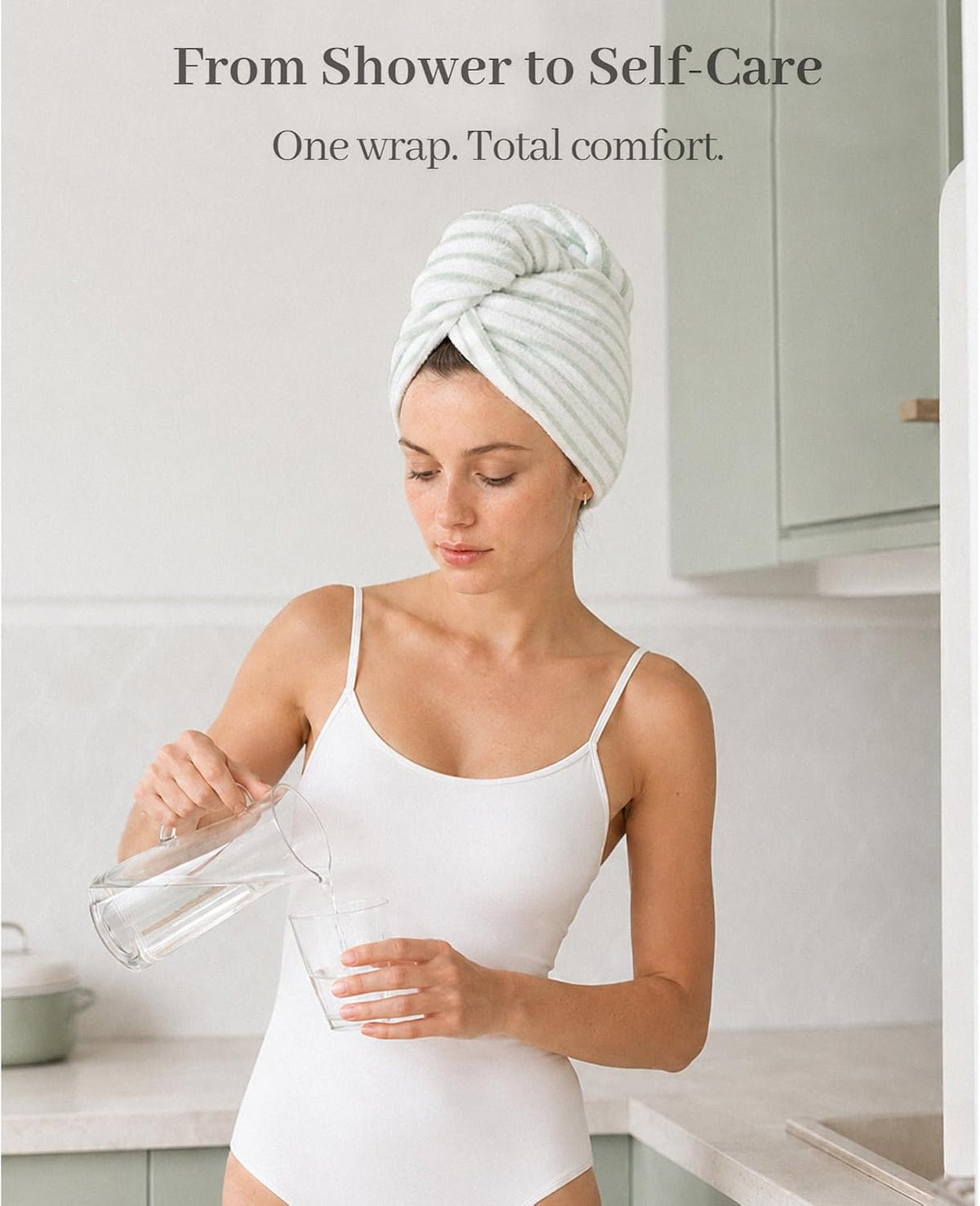 A light green and white striped hair towel wrap twisted into a turban on a woman's head. She wears a white camisole and pours water in a soft, neutral kitchen setting. Text reads 'From Shower to Self-Care. One wrap. Total comfort.'