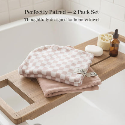 Two folded hair towel wraps on a wooden bath tray. The top towel has a pink and white checkerboard pattern; the bottom is solid blush pink. Small brand labels are visible on each. A bar of soap, loofah, and amber dropper bottle sit nearby.