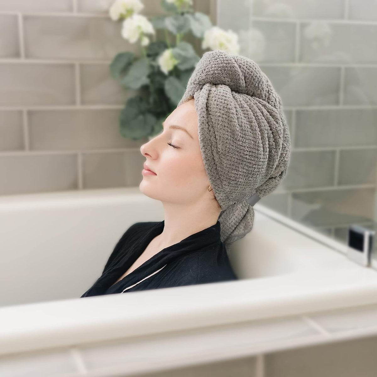 Woman relaxing in a bathtub with eyes closed, wearing a gray textured microfiber hair towel wrapped in a turban style on her head. She wears a dark robe in a tiled bathroom setting.