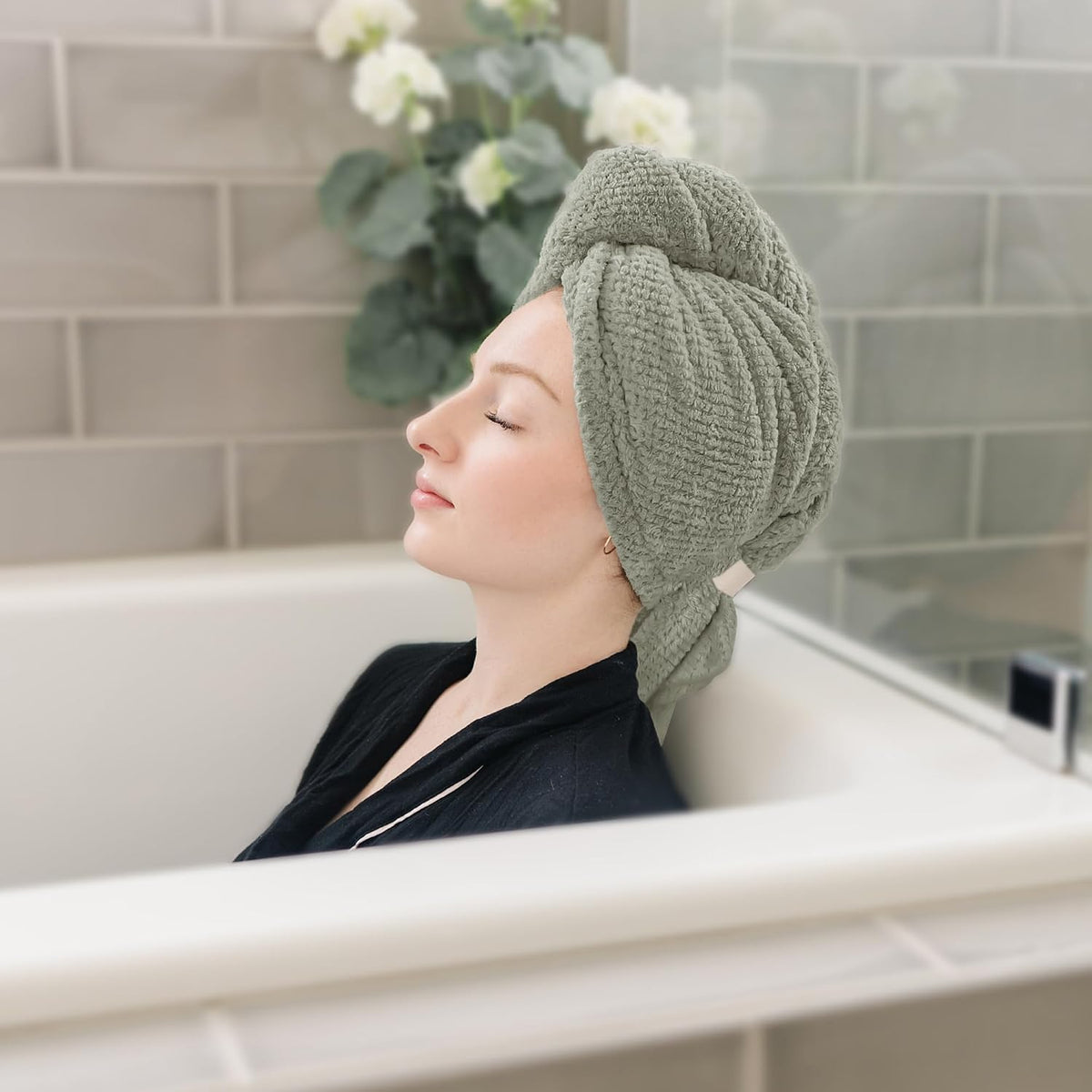 A woman with eyes closed relaxes in a bathtub wearing a sage green textured hair towel wrapped in a turban style on her head. The towel has a waffle-knit texture and a small light-colored tab closure at the side.