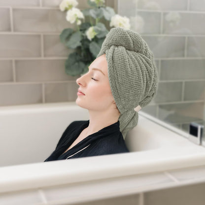 A woman with eyes closed relaxes in a bathtub wearing a sage green textured hair towel wrapped in a turban style on her head. The towel has a waffle-knit texture and a small light-colored tab closure at the side.