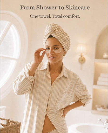 Woman in a bathroom wearing a beige houndstooth-patterned hair towel wrap twisted on her head, applying an under-eye patch. She wears a striped button-up shirt. Text reads 'From Shower to Skincare. One towel. Total comfort.'