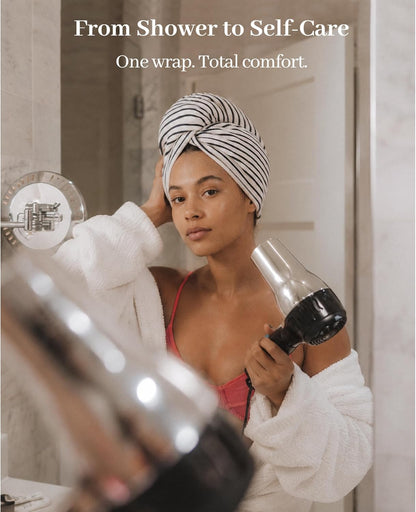 Woman in a white robe wearing a black-and-white striped hair towel turban, holding a hair dryer while looking in a bathroom mirror. Text reads 'From Shower to Self-Care. One wrap. Total comfort.'