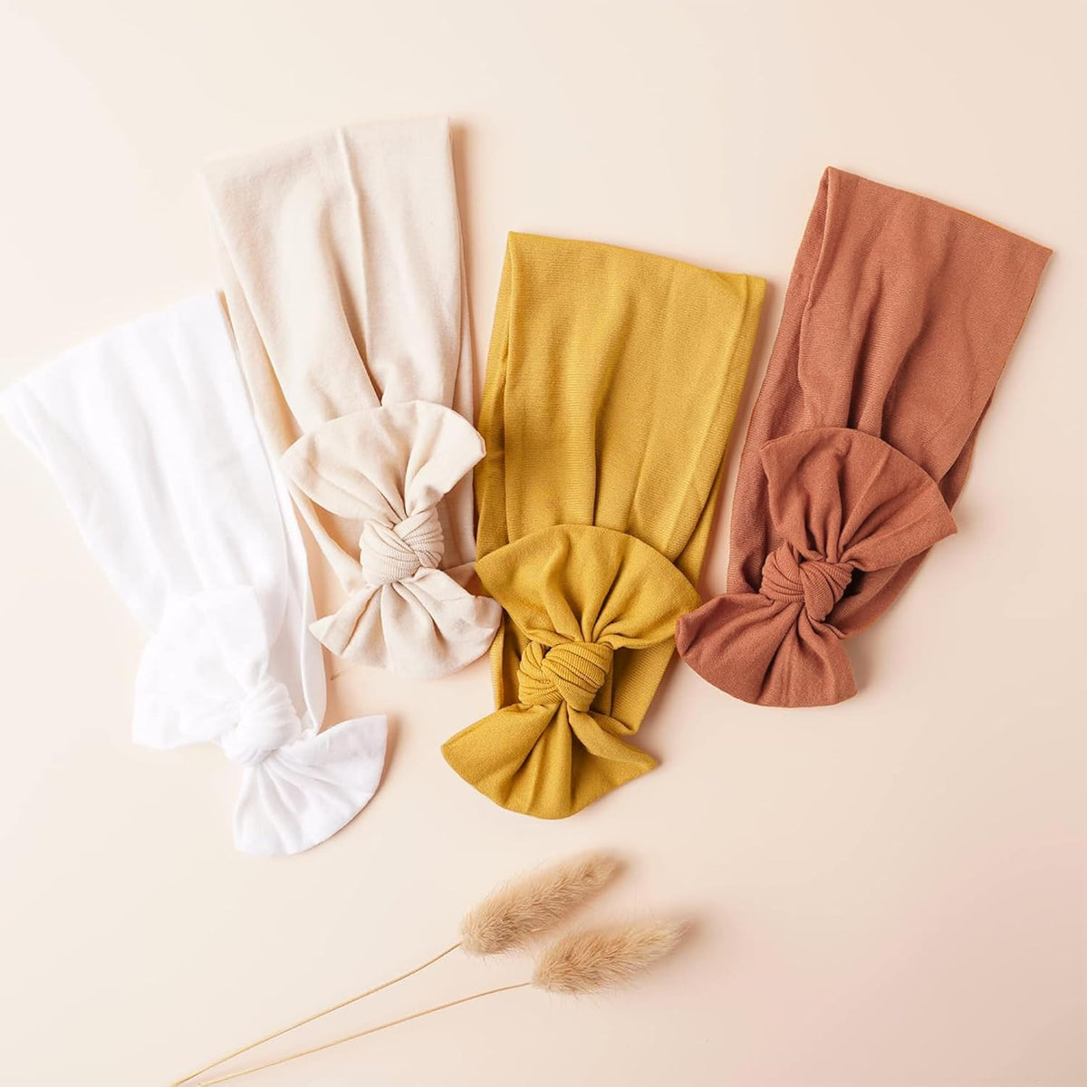 Four fabric headbands with knotted bows laid flat on a cream surface, in white, ivory, mustard yellow, and terracotta. Each band is wide and stretchy with a large tied bow at one end. A dried grass stem is placed nearby.